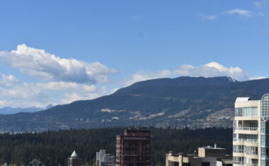 A birds-eye image of the Vancouver skyline. In the foreground, tall office and condo buildings; behind, the evergreens of Stanley Park. In the distance, you can see the neighbourhoods of North Vancouver; further still, snow-capped mountains. It's a sunny day, with billowing clouds.