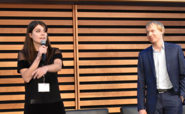 Samantha Moore standing on a stage holding a microphone with her right hand and then point to the right with her left hand. Behind her stands Jack McCormick with his arms behind his back. On the other side of her is Patrick Losier.