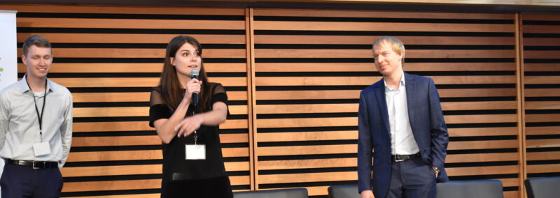 Samantha Moore standing on a stage holding a microphone with her right hand and then point to the right with her left hand. Behind her stands Jack McCormick with his arms behind his back. On the other side of her is Patrick Losier.