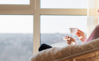 older woman sitting in a wide chair while looking out of a bright big window with a cup of tea in hand