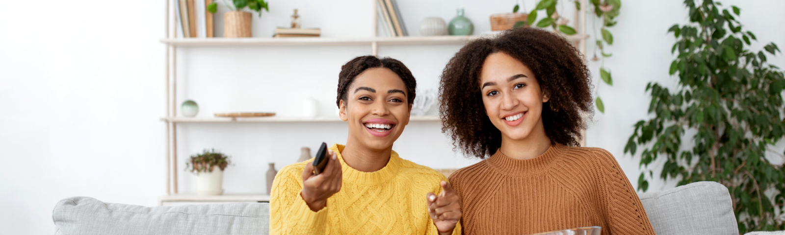 Two women laughing, eating a snack and holding a remote control, whilst watching television.