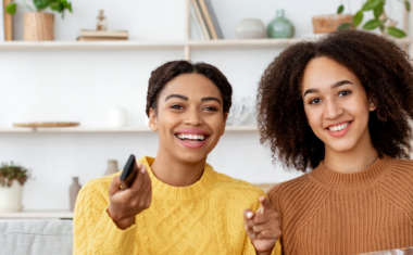 Two women laughing, eating a snack and holding a remote control, whilst watching television.