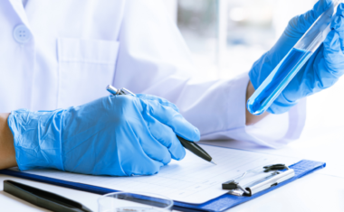 image is of a medical researcher wearing medical gloves, holding a vile and recording information onto a clipboard inside a laboratory.