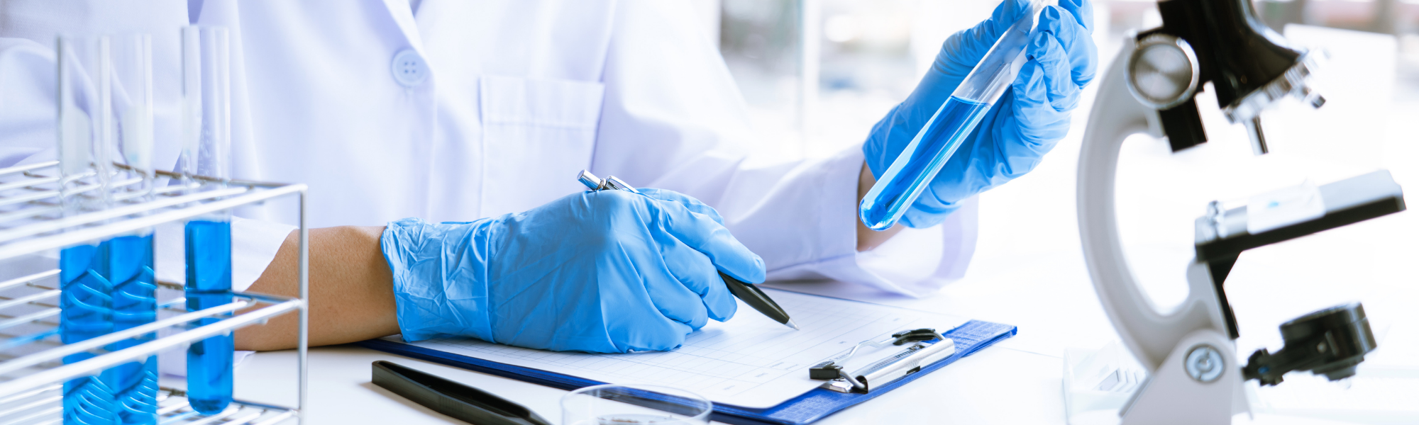 image is of a medical researcher wearing medical gloves, holding a vile and recording information onto a clipboard inside a laboratory.