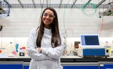 Woman wearing labcoat and goggles standing in front of a research desk.