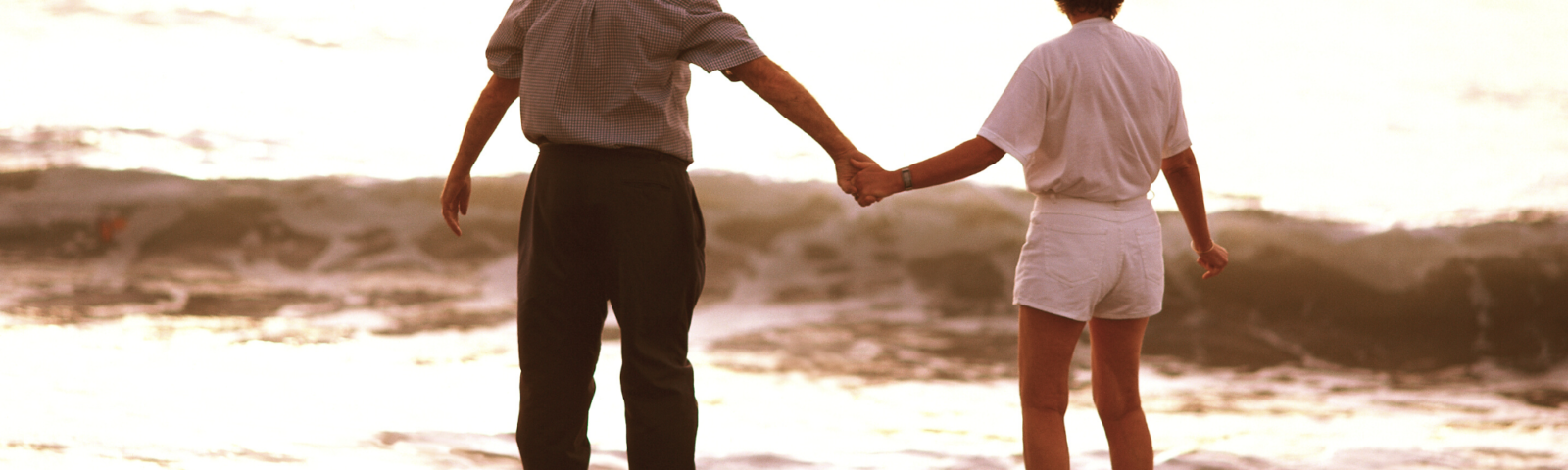 Image is of mature couple holding hands standing at the inlet of a beach. They are in the distance and are seen from behind.