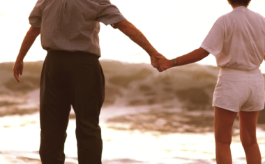 Image is of mature couple holding hands standing at the inlet of a beach. They are in the distance and are seen from behind.