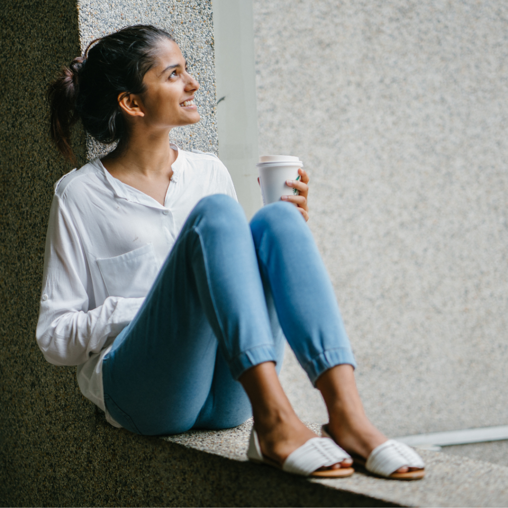 Image is of a woman sitting outside with a to-go beverage looking up at her surroundings and smiling