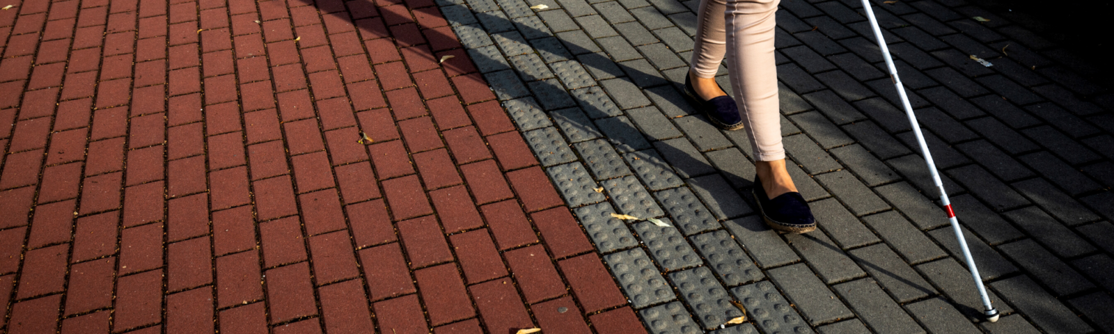 Image is of a woman's feet walking on across interlocking on a street using a white cane