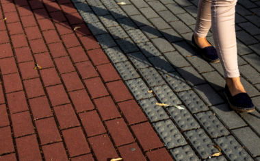 Image is of a woman's feet walking on across interlocking on a street using a white cane