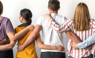 Image is of a group of people with their arms over each other's shoulders. Their backs are facing forward.