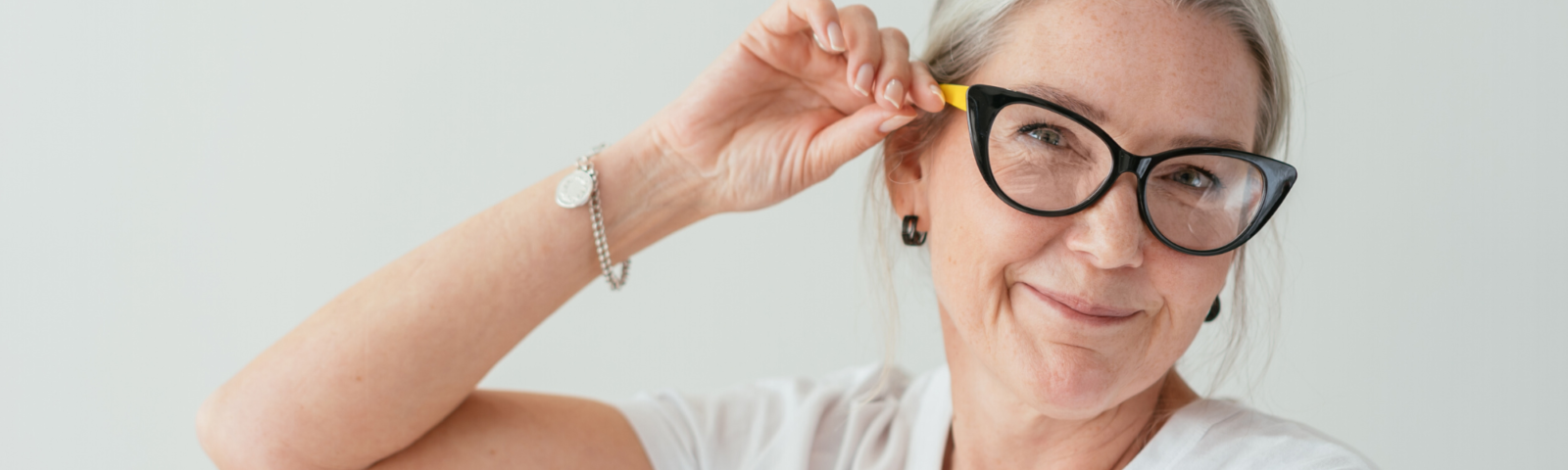 Image is of a mature woman wearing glasses and touching the frame whilst smiling.