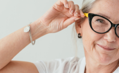 Image is of a mature woman wearing glasses and touching the frame whilst smiling.