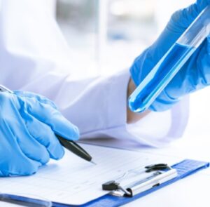Image is of hands holding a medical vile of liquid and recording notes whilst wearing medical gloves.