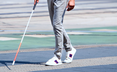 Image is of a legs using a white cane walking across a road.