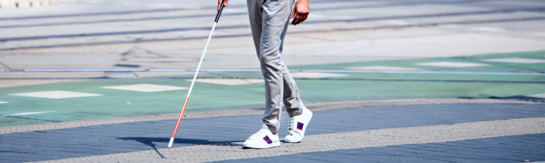 Image is of a legs using a white cane walking across a road.