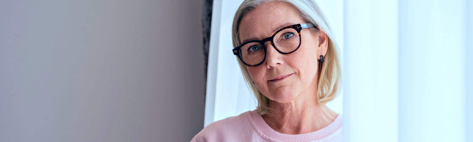 Image is of a mature woman leaning against an open window. She is wearing eyeglasses.
