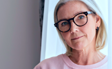 Image is of a mature woman leaning against an open window. She is wearing eyeglasses.