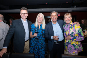 Image is of 4 event attendees, smiling for the camera while standing and holding beverages.