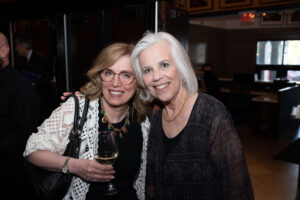 Image is of 2 event attendees smiling for the camera, closely to one and other, while holding beverages.