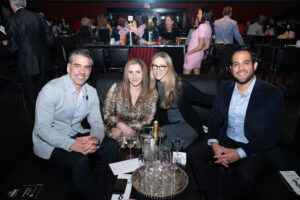 Image is of event attendees in a booth at the comic vision event. They are smiling for the camera with bottle service and glassware in front of them.