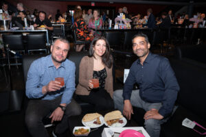 Image is of three event attendees seated, smiling for the camera, and holding beverages at Comic Vision.