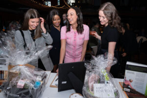 Image is of a group of young women smiling and laughing while particpating in the auction. They are standing around the auction item bidding cards.