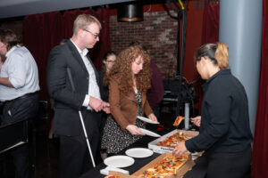 Image is of 2 event attendees being served at the food station by a waiter.