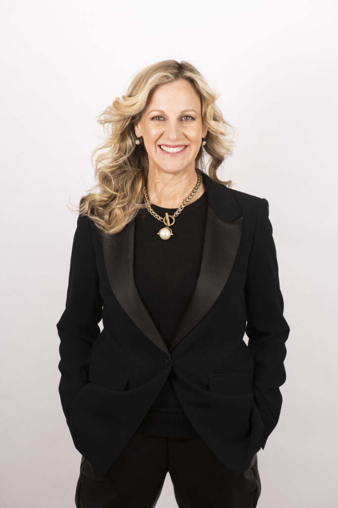 Image is of Jennifer Jones. She is smiling wearing an all-black suit with necklaces.