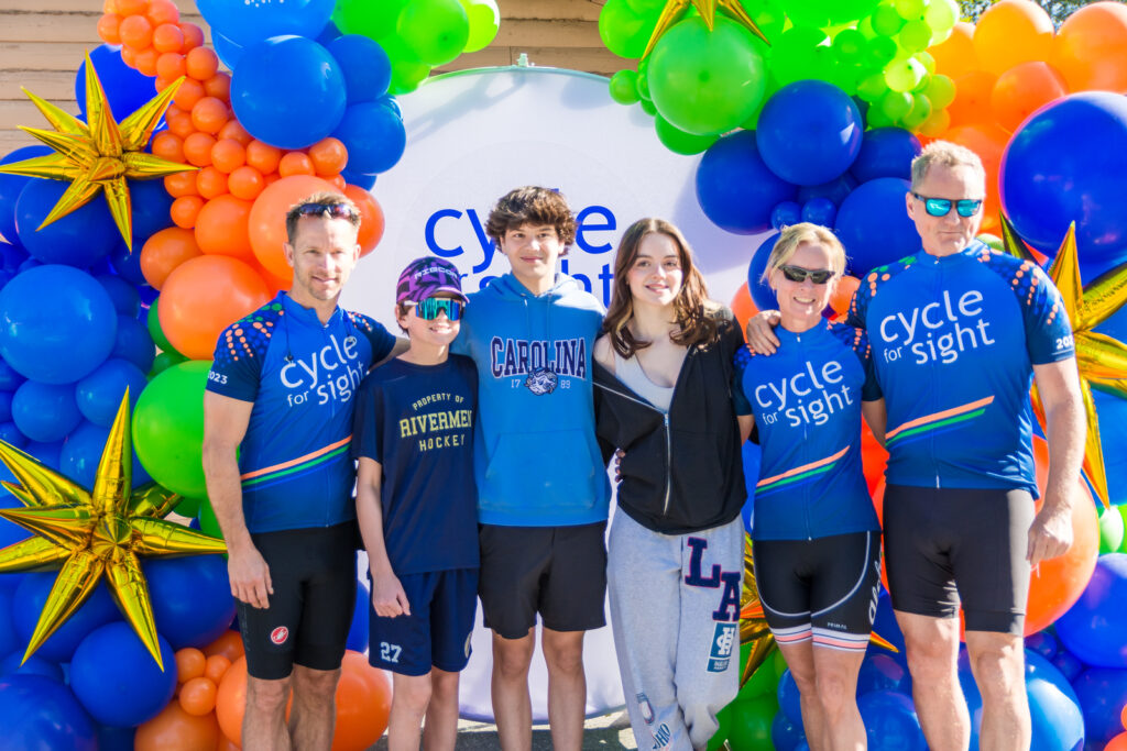 Image is of a group of youngsters and parents smiling in front of Cycle for Sight signage.