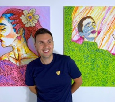 Image of Adam Lancia wearing a dark blue shirt with a yellow heart on the right side standing in front of two of his paintings.