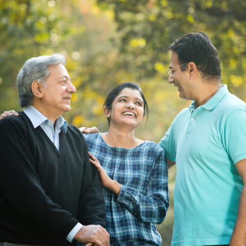 Three people stand together outdoors smiling warmly at each other. An older man with grey hair stands on the left, a young woman in a plaid shirt is in the centre, and a middle-aged man in a teal polo shirt stands on the right. They appear to be a family enjoying time together in a natural outdoor setting with green trees in the background.