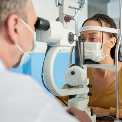 A female patient wearing a face mask sits at a slit lamp while a male doctor in a white coat and face mask conducts an eye examination in a clinical setting